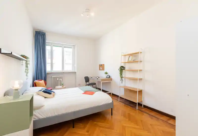 Dormitorio luminoso y bien decorado con cama doble, mesita con lámpara, escritorio junto a la ventana y estantería abierta. Suelo de parquet cálido y textiles suaves crean un ambiente acogedor.
