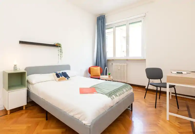 Dormitorio moderno y luminoso con cama doble, mesita, textiles acogedores y un escritorio junto a la ventana. La luz natural y el suelo de parquet aportan calidez.