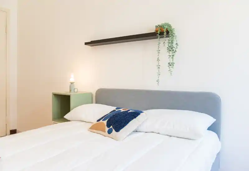 Dormitorio moderno y luminoso con cama bien hecha, cojín decorativo, lámpara de mesita y planta colgante — estilo minimalista y limpio, perfecto para el anuncio.