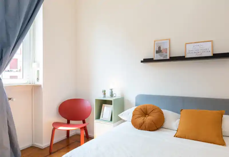 Camera da letto luminosa e curata, con letto confortevole, cuscini dai toni caldi e una sedia decorativa vicino alla finestra — perfetta per annunci che puntano su comfort e luce.