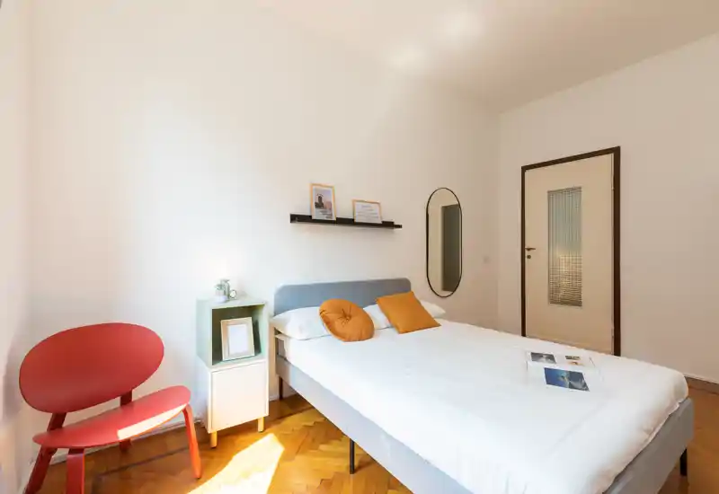 Dormitorio moderno y luminoso con cama doble, cojines cálidos y luz natural; decoración sencilla con estante, espejo y una silla roja.