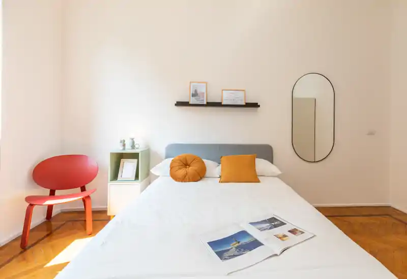 Dormitorio luminoso y minimalista con una cama doble bien hecha, cojines color mostaza, una repisa decorativa y un espejo ovalado; suelo de parqué limpio y luz natural que crean un ambiente acogedor.