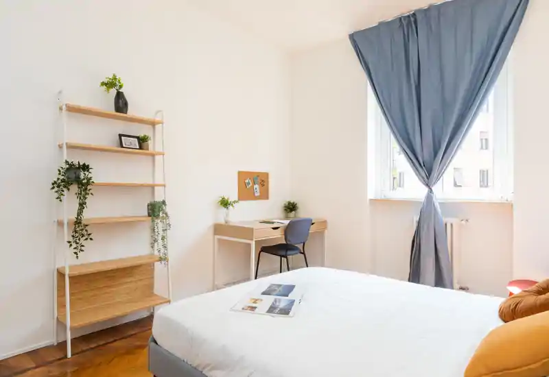 Camera da letto luminosa e ben arredata con letto bianco, cuscini morbidi e una scrivania accanto a una finestra ampia che lascia entrare molta luce naturale.