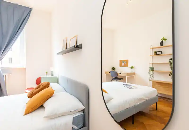 Dormitorio moderno y luminoso con una cómoda cama doble y cojines coloridos, luz natural desde la ventana y un espejo grande que aporta profundidad y estilo.