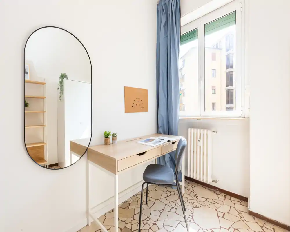 Angolo lavoro luminoso e minimal con scrivania in legno, sedia imbottita, specchio grande e luce naturale da una finestra: perfetto per smart working o studio.