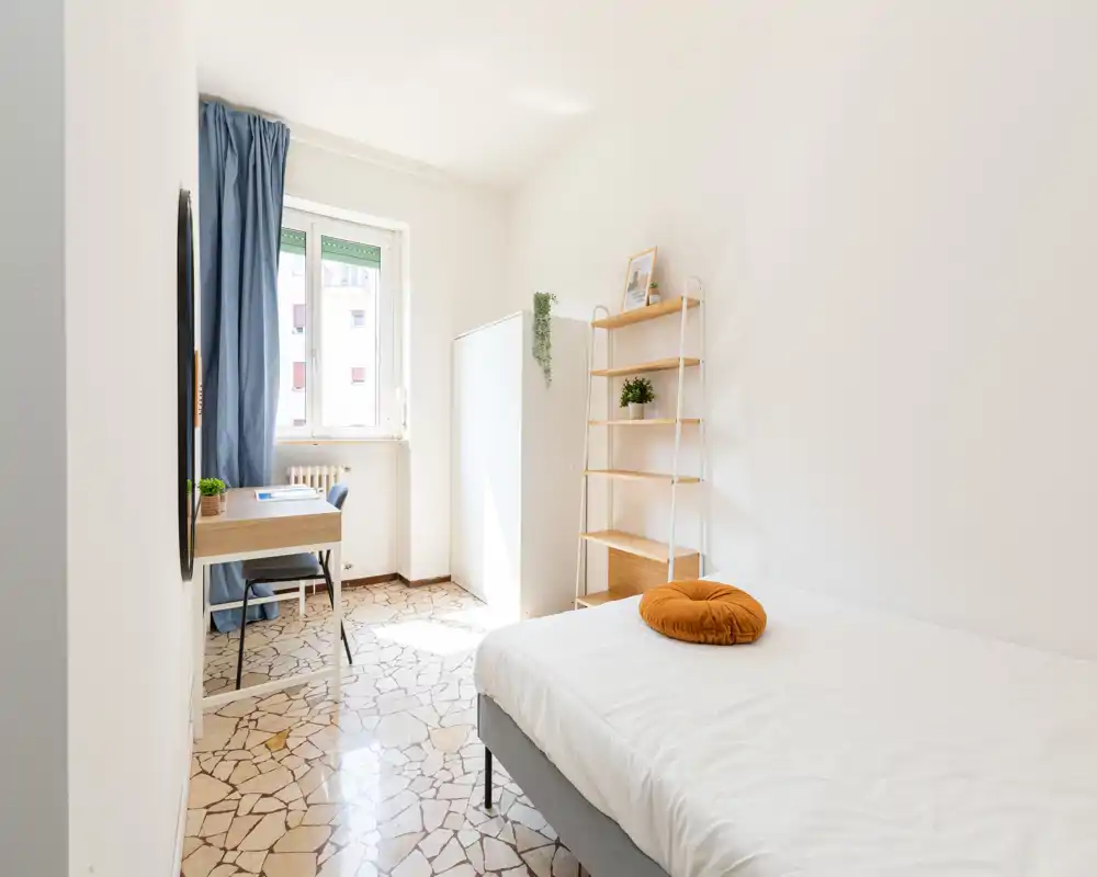 Camera da letto luminosa e minimale con letto ordinato, una mensola a gradini in legno, scrivania vicino alla finestra e un cuscino accentuato: perfetta per annunci che valorizzano la luce naturale e l’arredo pulito.