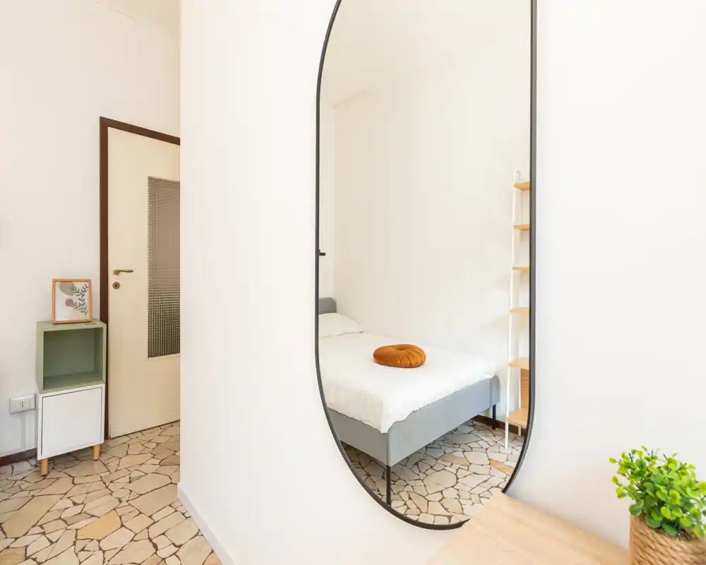 Camera da letto luminosa e minimale con letto singolo grigio, biancheria bianca e un cuscino rotondo decorativo; uno specchio alto riflette lo spazio e ne aumenta la profondità.