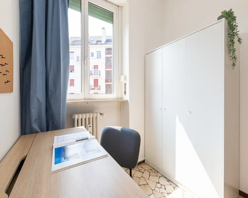 Piccola stanza luminosa con scrivania davanti alla finestra, una sedia imbottita e un armadio bianco — ideale come postazione di lavoro o angolo studio per una persona.