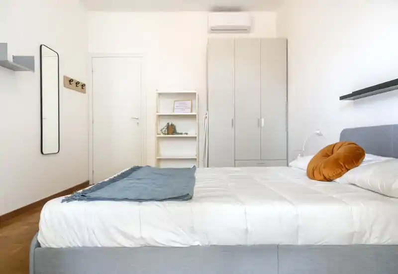 Camera da letto moderna e luminosa con letto matrimoniale ben rifatto, testiera grigia e cuscino d'accento caldo. Layout minimale, ben illuminato, con guardaroba e mensole ideale per annunci di affitto.