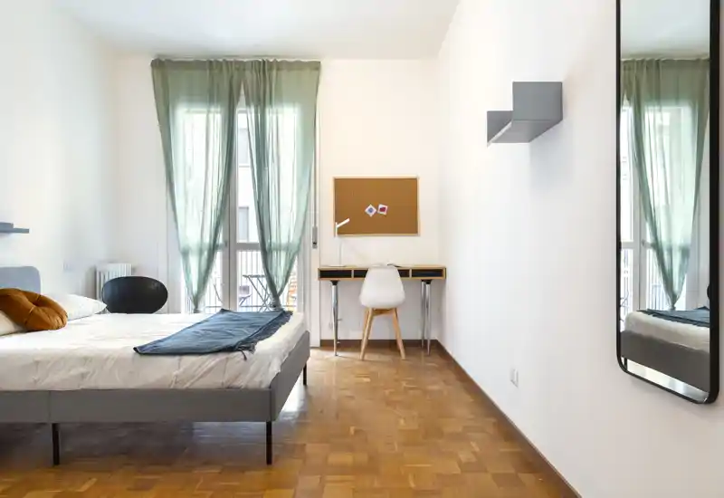 Camera da letto luminosa e minimalista con letto matrimoniale, cuscini decorativi e una scrivania vicino a una grande finestra con tende verdi leggere; pavimento in parquet pulito e uno specchio a parete aumentano la sensazione di spazio.