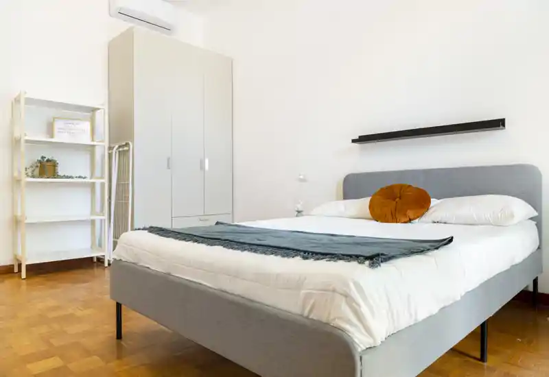 Camera da letto luminosa e moderna con letto matrimoniale ben rifatto, cuscino decorativo e scaffalatura semplice; pavimento in parquet e linee pulite creano un ambiente accogliente.