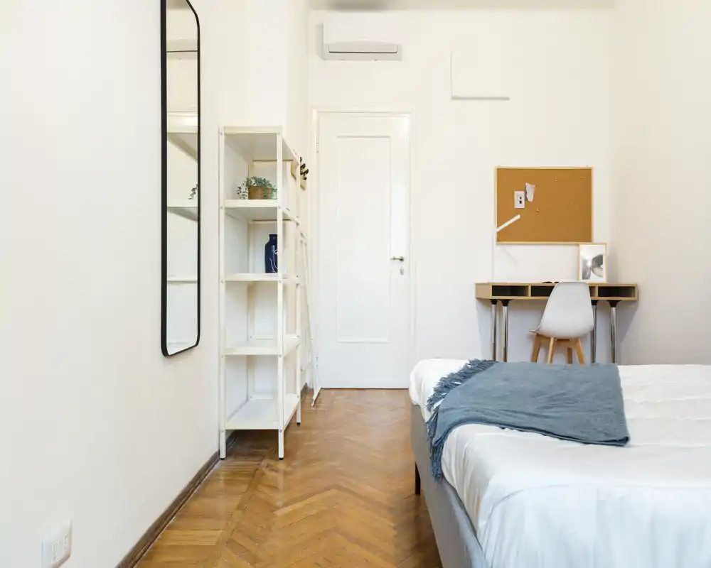 Dormitorio luminoso y minimalista con cama bien hecha, pequeño escritorio y estantería; suelo de madera en espiga y decoración neutra que transmiten calma.