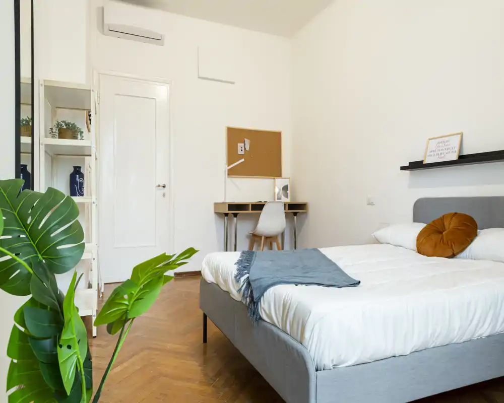 Dormitorio luminoso y moderno con cama tapizada gris, ropa blanca, cojín redondo decorativo y manta azul; suelo de parqué y plantas que aportan calidez y estilo.