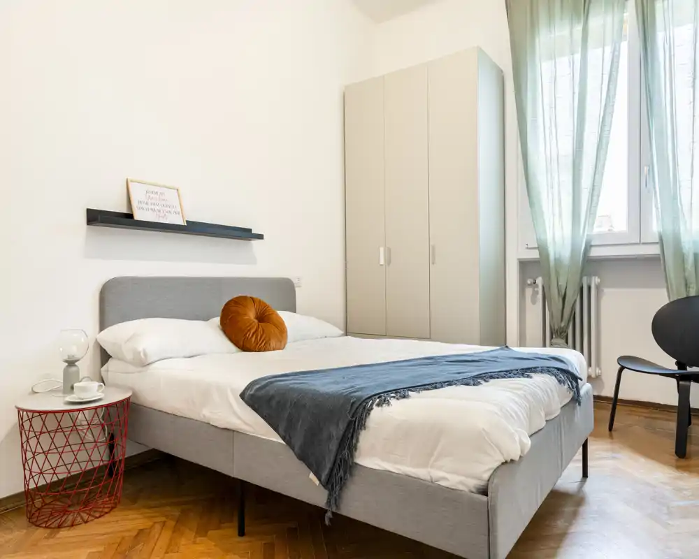 Dormitorio luminoso y moderno con cama doble bien hecha, armario y luz natural procedente de una ventana amplia. Suelo de madera y decoración sencilla y cuidada.