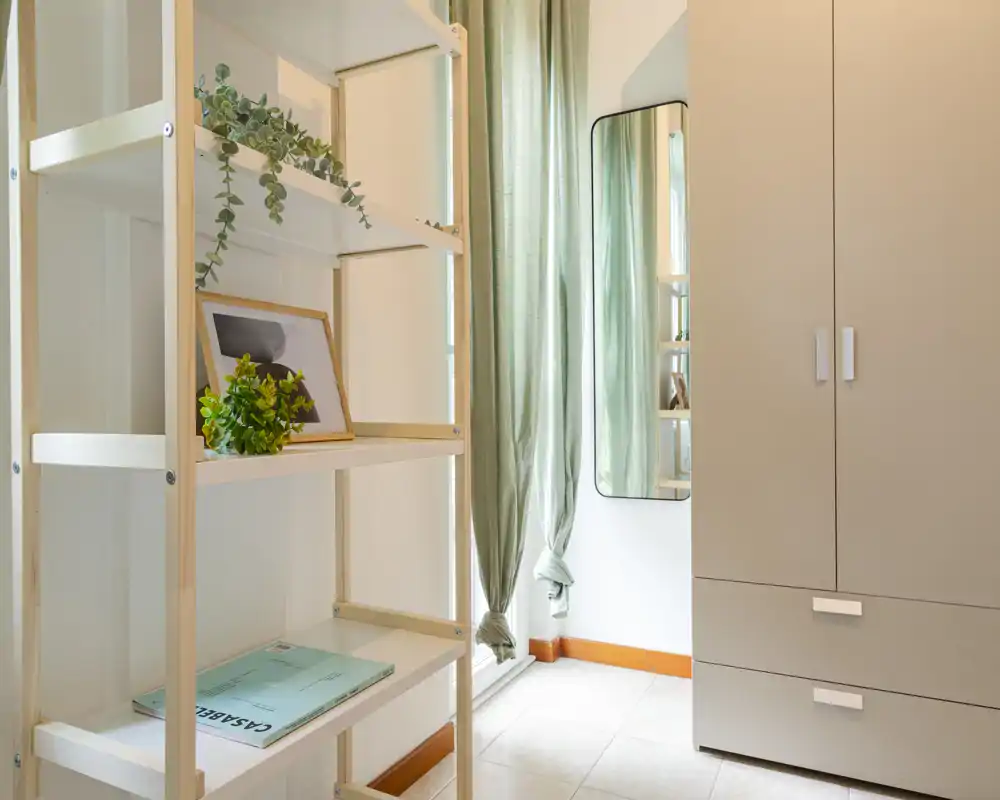 Rincón de dormitorio pequeño y luminoso con armario, estantería y espejo de cuerpo entero junto a una ventana con cortinas verdes — limpio, minimalista y bien iluminado.