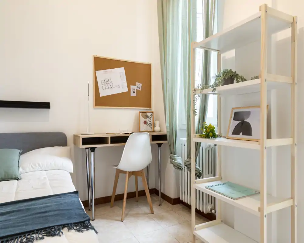Habitación luminosa y moderna con una cama individual cómoda, un pequeño escritorio junto a la ventana y estanterías abiertas con plantas y decoración — ideal para estudiante o una sola persona.