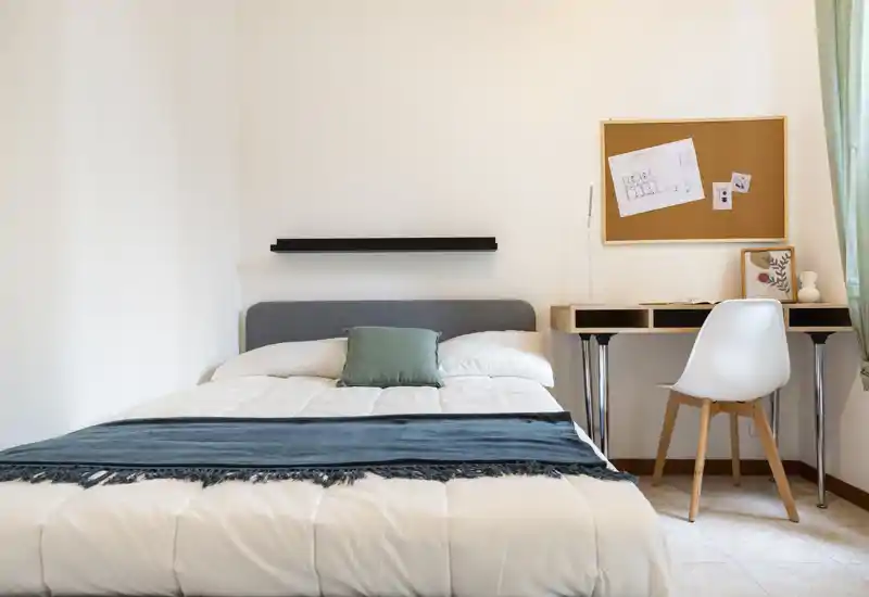 Camera da letto luminosa e minimal con un ampio letto, biancheria bianca e cuscino turchese; sulla destra una scrivania con sedia sotto una bacheca e luce filtrata dalla finestra.