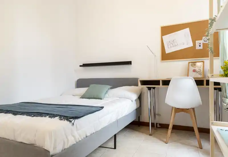 Camera da letto compatta e luminosa con letto comodo, biancheria morbida, scrivania e bacheca: ideale per uno studente o un professionista single.