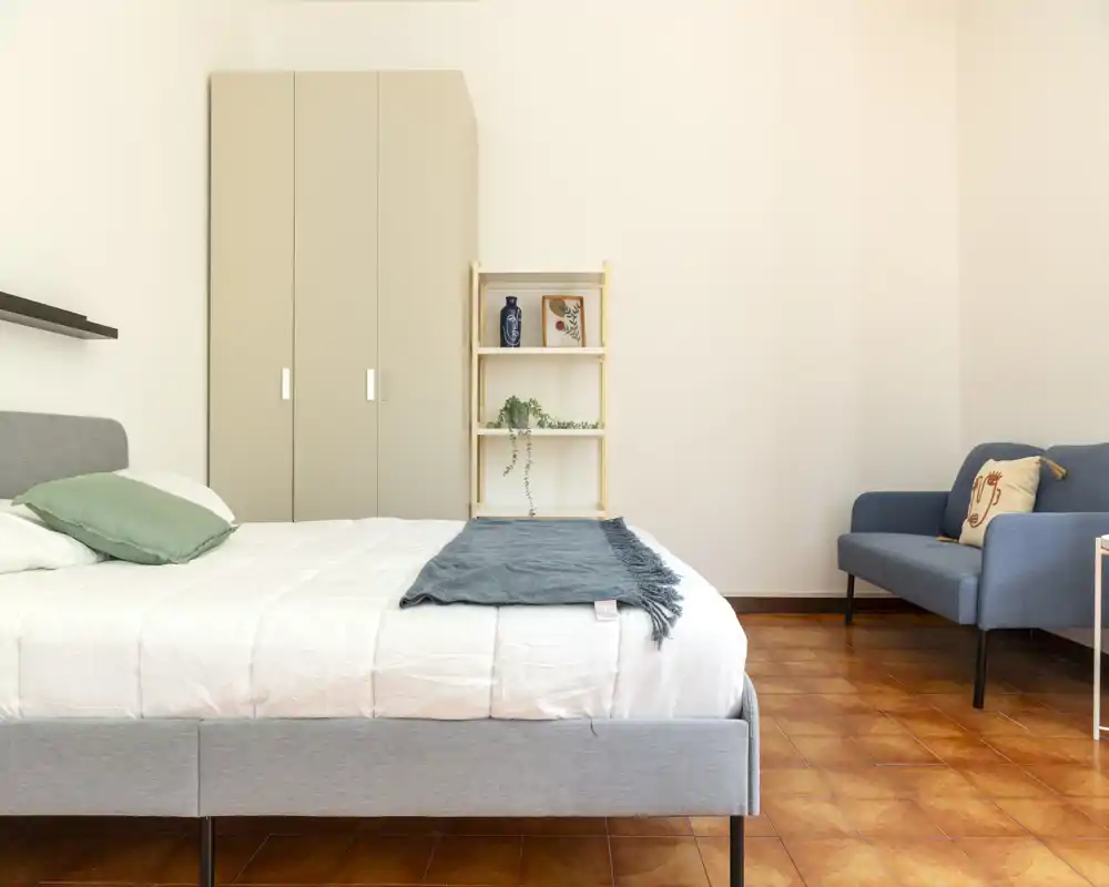 Dormitorio luminoso y moderno con cama bien hecha, textiles suaves y un pequeño rincón de estar — ideal para anuncios que destaquen comodidad y diseño limpio.