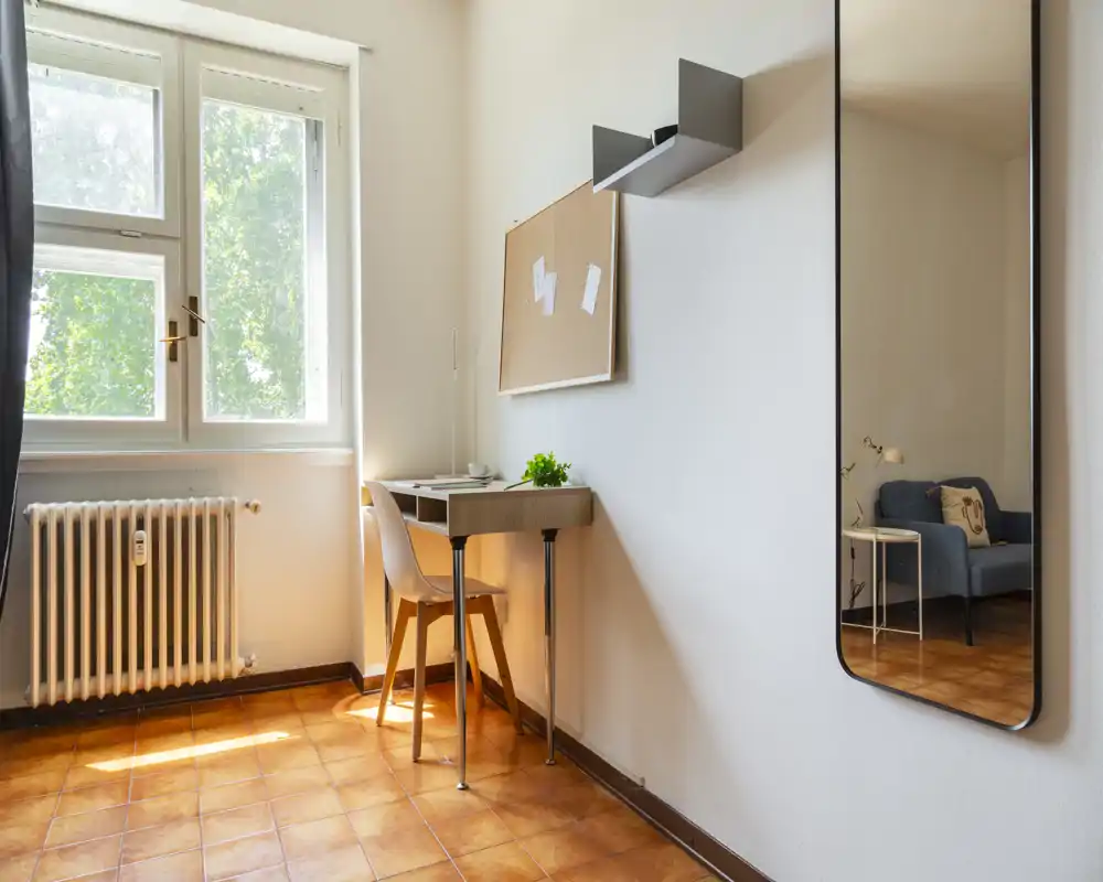 Área de estar luminosa y minimalista con ventana que aporta luz natural, un pequeño escritorio y silla, y un espejo que refleja un sillón acogedor — ideal como rincón funcional de vida o trabajo.