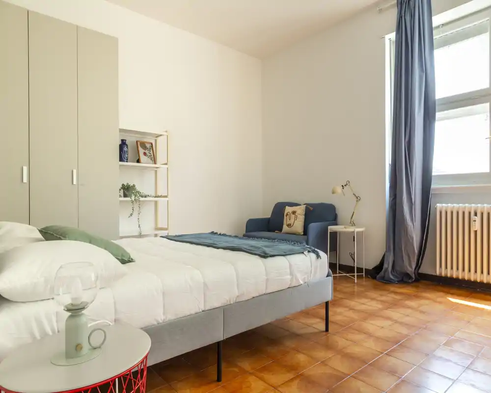 Dormitorio luminoso y moderno con cama doble bien hecha, pequeño sillón, armario y luz natural procedente de una gran ventana — estilo minimalista y relajante ideal para un anuncio.