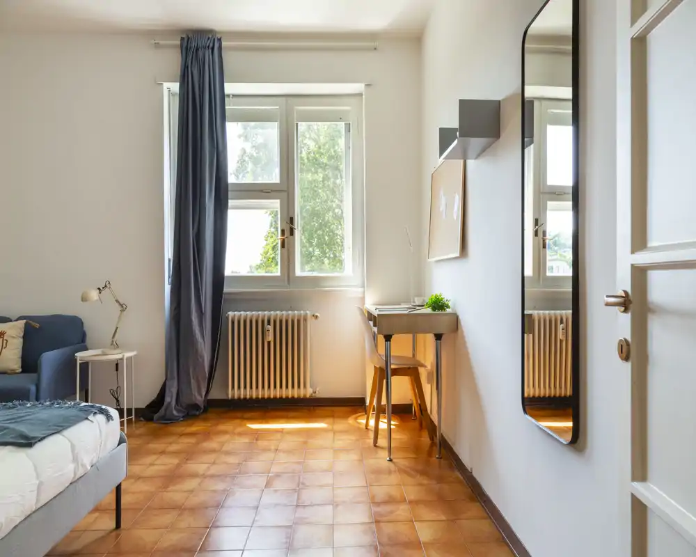 Dormitorio luminoso y acogedor con luz natural de una gran ventana, cama visible en primer plano y un pequeño espacio de trabajo junto a la pared.