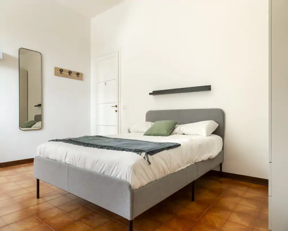 Dormitorio luminoso y minimalista con una cama doble bien hecha, ropa de cama suave y una repisa sobre el cabecero. El suelo de terracota y las paredes neutras crean un ambiente tranquilo y moderno, perfecto para alquileres de corta estancia.