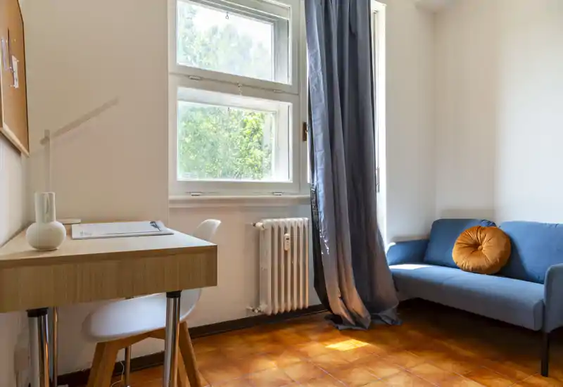 Espace salon lumineux et soigné avec un petit canapé, un coussin décoratif rond et un bureau près de la fenêtre — idéal pour se détendre ou travailler.