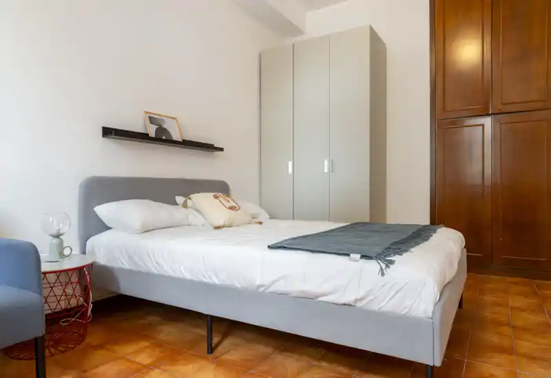 Chambre lumineuse au style épuré avec lit rembourré gris, linge de lit blanc et plaid, armoire et sol carrelé chaud — confortable et bien éclairée.
