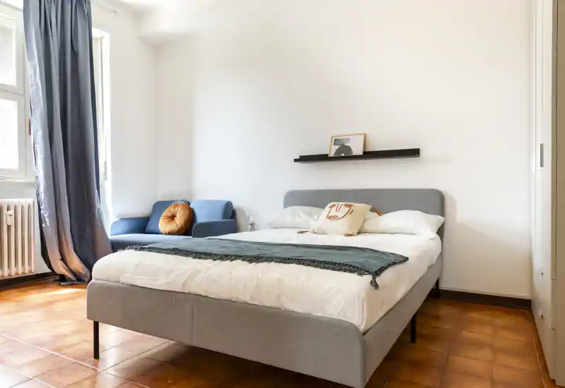 Chambre lumineuse et soignée avec un lit double entièrement visible, du linge de lit doux et un petit coin salon près de la fenêtre — bien éclairée et décorée sobrement pour le confort.