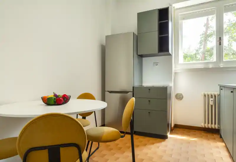 Small, bright kitchen corner with a round dining table, mustard-yellow chairs, fridge and modern storage units; natural light from a window creates a welcoming atmosphere.