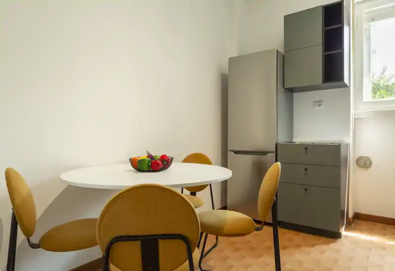 Bright, compact dining area with a round white table, mustard upholstered chairs and a fruit bowl; neat layout next to kitchen storage and a window providing natural light.