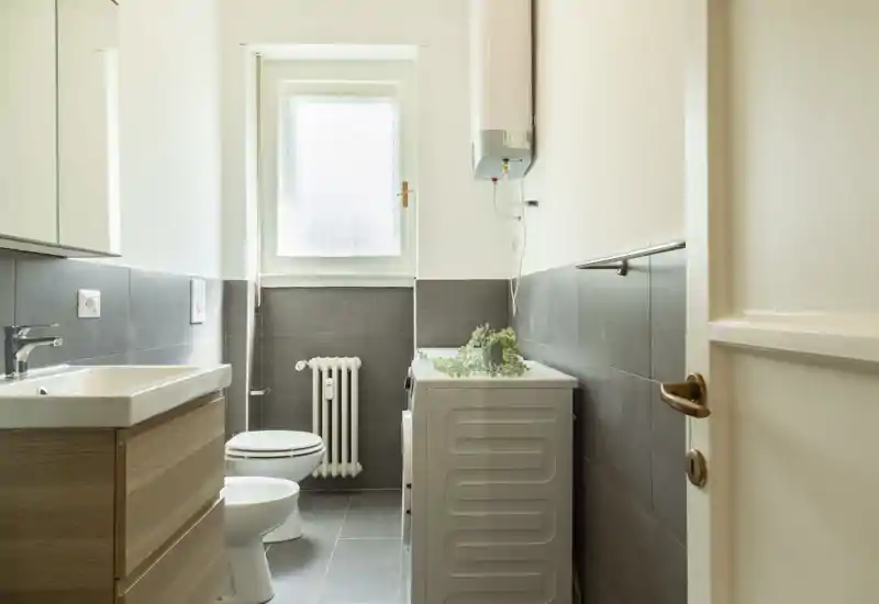Bright, modern bathroom with a sink vanity, toilet and a washing machine by the window. Neutral tiles and natural light create a clean, functional space.