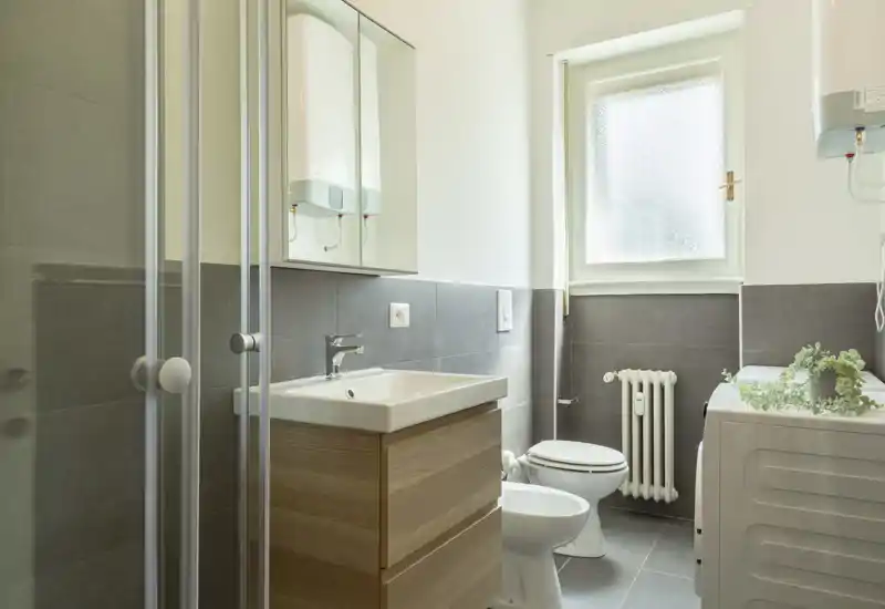 Bright, modern bathroom with a shower cabin, sink with vanity, toilet and bidet. Neutral gray tiles and a frosted window provide privacy and natural light.