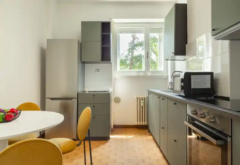 Bright modern kitchen with matte gray cabinetry, stainless refrigerator, built-in oven and a round dining table with warm yellow chairs — a compact, well-lit space ideal for a small household.