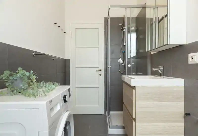 Compact modern bathroom with a glass shower enclosure, sink with wooden vanity, and a washing machine — clean neutral tiles and bright natural light.