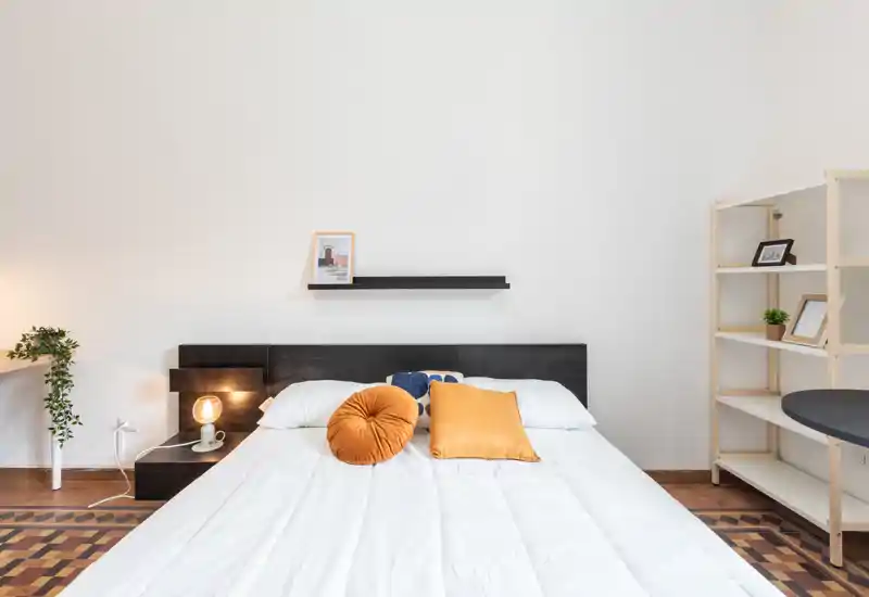 Dormitorio moderno y luminoso con cama grande, cojines decorativos cálidos y estanterías minimalistas; líneas limpias y luz suave que crean un espacio acogedor.