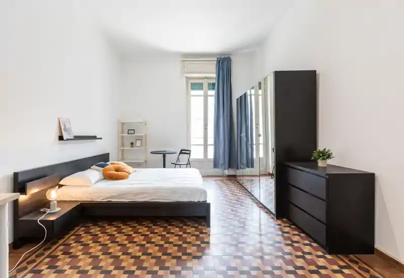 Dormitorio moderno y luminoso con amplia cama doble, suelo de parqué con patrón y armario con espejo; la luz natural inunda la habitación creando un espacio aireado y estiloso.