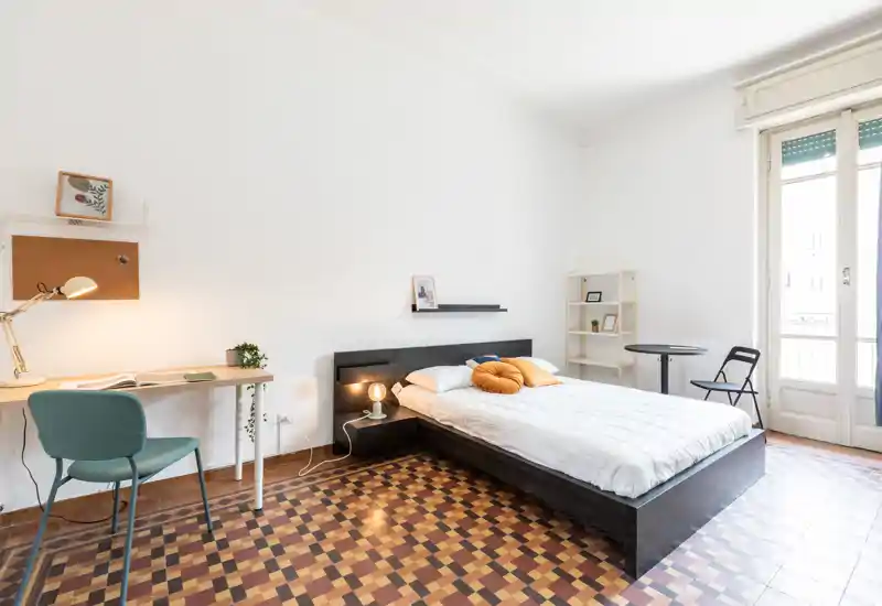 Dormitorio luminoso y moderno con cama baja doble, cojines decorativos, lámpara de mesita, escritorio y una gran ventana-balconcillo que aporta mucha luz natural.