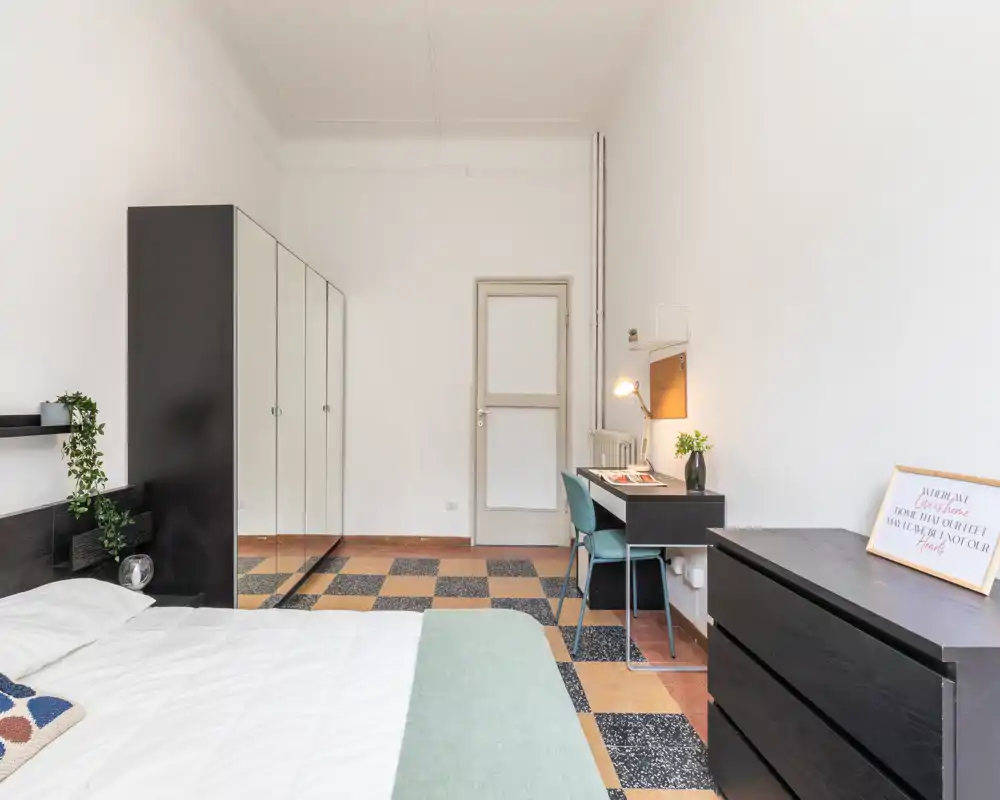 Dormitorio luminoso y minimalista con cama grande, armario con espejo y pequeña zona de escritorio — espacio limpio y despejado con suelo de terrazo con patrón.