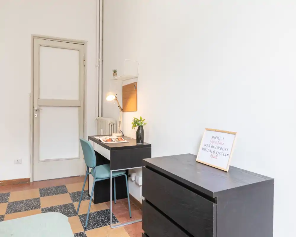 Rincón de dormitorio luminoso y minimalista con un pequeño escritorio y cómoda; se ve parte de la cama en la esquina inferior izquierda. El espacio está amueblado de forma sencilla con lámpara de mesa, silla y planta decorativa, ideal para un cuarto de alquiler compacto.