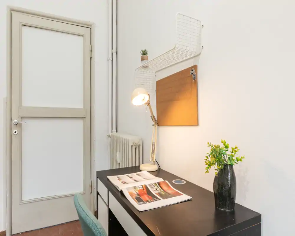 Pequeño espacio de trabajo con escritorio negro, lámpara ajustable, corcho y una planta decorativa — bien organizado para estudiar o teletrabajar.