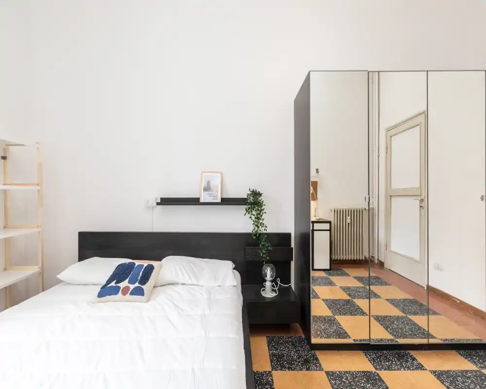 Dormitorio luminoso y minimalista con una cama grande, ropa de cama blanca, cabecero oscuro y un armario con espejos que añade profundidad. Estilo limpio con planta y estantería sencilla.