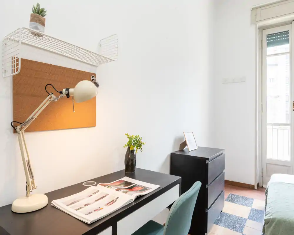 Dormitorio con zona de trabajo ordenada: escritorio oscuro con lámpara y silla junto a una cama parcialmente visible y puerta al balcón que aporta luz natural.