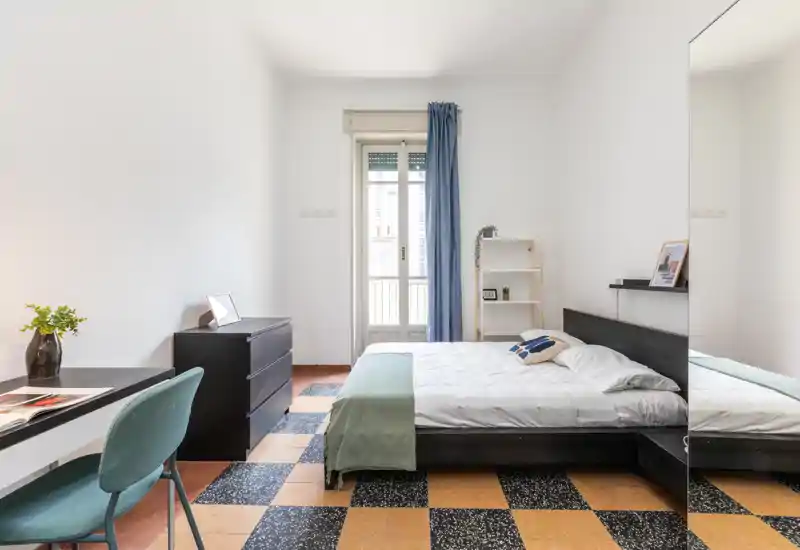 Dormitorio luminoso y minimalista con cama doble, luz natural desde la puerta balcón y mobiliario sencillo y moderno como escritorio y cómoda.