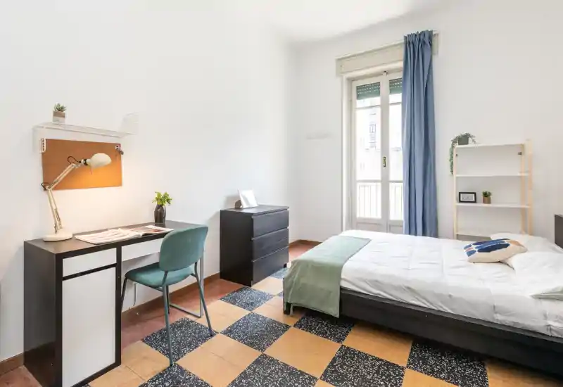 Dormitorio luminoso y bien presentado con cama doble, escritorio y estantería. La luz natural entra por la puerta al balcón y los textiles aportan una sensación acogedora.