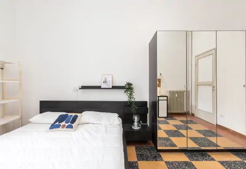 Dormitorio luminoso y minimalista con una cama grande, ropa de cama blanca, cabecero oscuro y un armario con espejos que añade profundidad. Estilo limpio con planta y estantería sencilla.