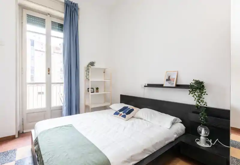 Dormitorio luminoso y minimalista con cama doble, estantería junto a la cama y puerta al balcón que aporta mucha luz — estilo moderno y sencillo con textiles y plantas.