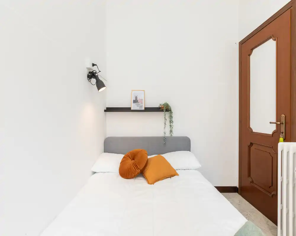 Chambre minimaliste et soignée avec lit double, coussins aux tons chauds, petite étagère murale et lampe murale — lumineuse et bien mise en scène pour location.