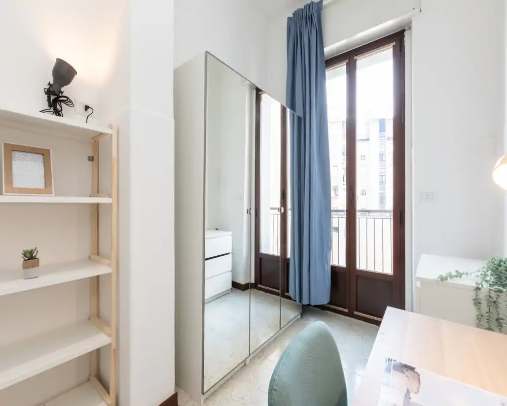 Coin de chambre lumineux et minimaliste avec une armoire miroir, un bureau et des portes-fenêtres qui laissent entrer la lumière naturelle. L'espace paraît frais et fonctionnel, avec un mobilier sobre et des rideaux bleu pâle.